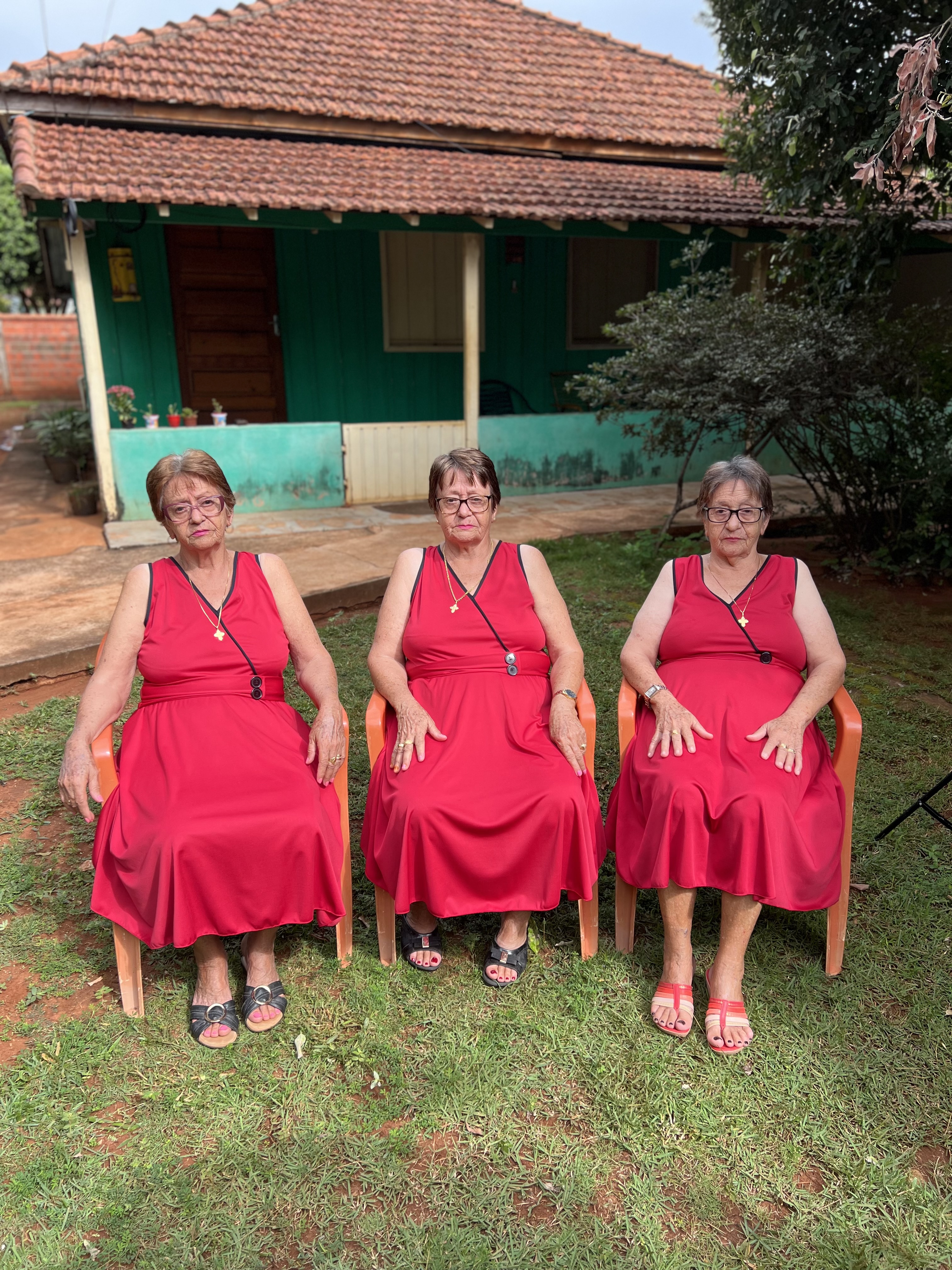 Três mulheres idosas sentadas em cadeiras de plástico laranja, vestindo vestidos vermelhos idênticos, posando em frente a uma casa simples com paredes verdes e telhado de telhas marrons. Elas estão lado a lado, no jardim, cercadas por vegetação, e com expressões calmas no rosto